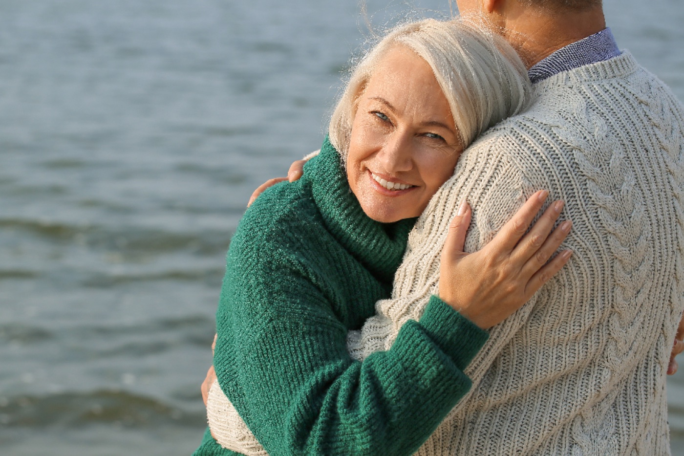 A smiling woman with silver hair stands outdoors in soft light, a scarf around her shoulders.